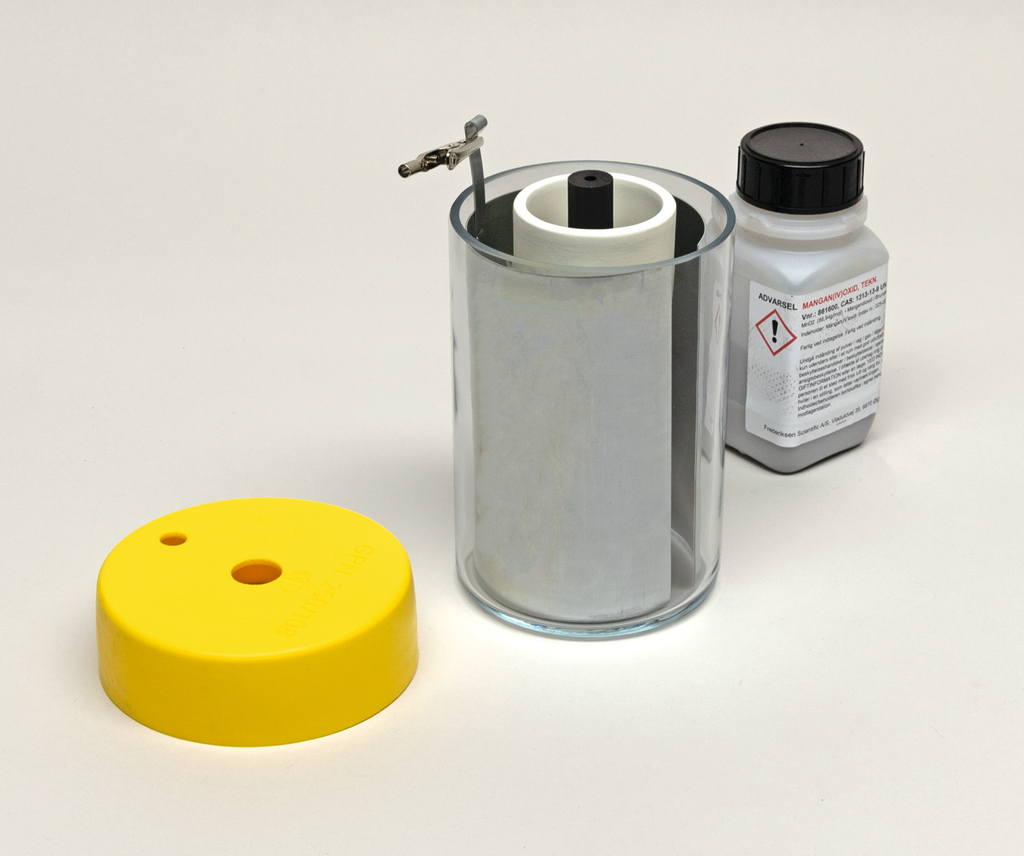 Das Bild zeigt ein transparentes Glasbehältnis mit einem weißen Innenteil, das offenbar für chemische Experimente verwendet wird. Daneben befindet sich eine gelbe Abdeckung und eine Flasche mit einer Warnaufkleber, die einen chemischen Stoff enthält.