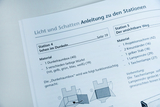 Das Bild zeigt eine Anleitung für experimentelle Stationen zum Thema Licht und Schatten im Bildungsbereich. Es beschreibt zwei Stationen, darunter Materialien und Anweisungen für Experimente, die das Sehen im Dunkeln und den unsichtbaren Weg thematisieren.