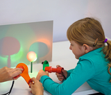 Das Bild zeigt ein Kind, das mit einem roten Taschenlampenähnlichen Gerät und einer orangefarbenen Linse experimentiert, während eine zweite Person mit einer orangefarbenen Linse arbeitet. Auf dem Tisch sind zwei Lichtquellen, die verschiedene Lichtfarben projizieren.