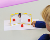 Das Bild zeigt einen Jungen, der mit einem gelben Spiegel und einem roten Würfel auf einem Tisch experimentiert. Die Spiegel reflektieren den Würfel, wodurch geometrische und optische Phänomene untersucht werden können.