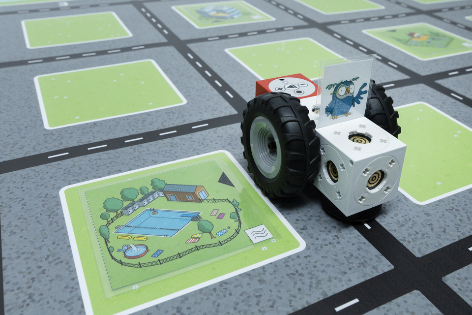 Detailbild von dem eXperiBot auf dem Spielplan f&uuml;r die Grundschule. Der eXperiBot steht neben dem Schwimmbad und kann &uuml;ber Programmierung zu anderen Orten auf dem Spielplan gelangen.