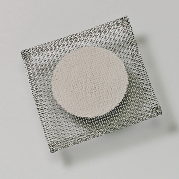 Das Bild zeigt eine runde, helle Probe, die auf einem feinmaschigen Drahtgitter platziert ist. Dieses Setup könnte für Experimente im Bildungsbereich verwendet werden, möglicherweise im Bereich der Chemie oder Materialwissenschaften.