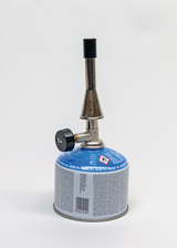 Das Bild zeigt einen Brenner mit einer blauen Gasflasche, der für experimentelle Zwecke verwendet wird. Der Brenner hat eine metallische Düse und ein Regelventil zur Einstellung der Flammenstärke.