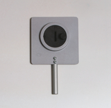 Das Bild zeigt ein Experimentiergerät, bestehend aus einer rechteckigen Platte mit einer runden Öffnung in der Mitte und einem drehbaren Regler. Es ist mit einem Metallstab ausgestattet, der als Halterung dient, und wird vermutlich zur Durchführung von physikalischen Experimenten verwendet.