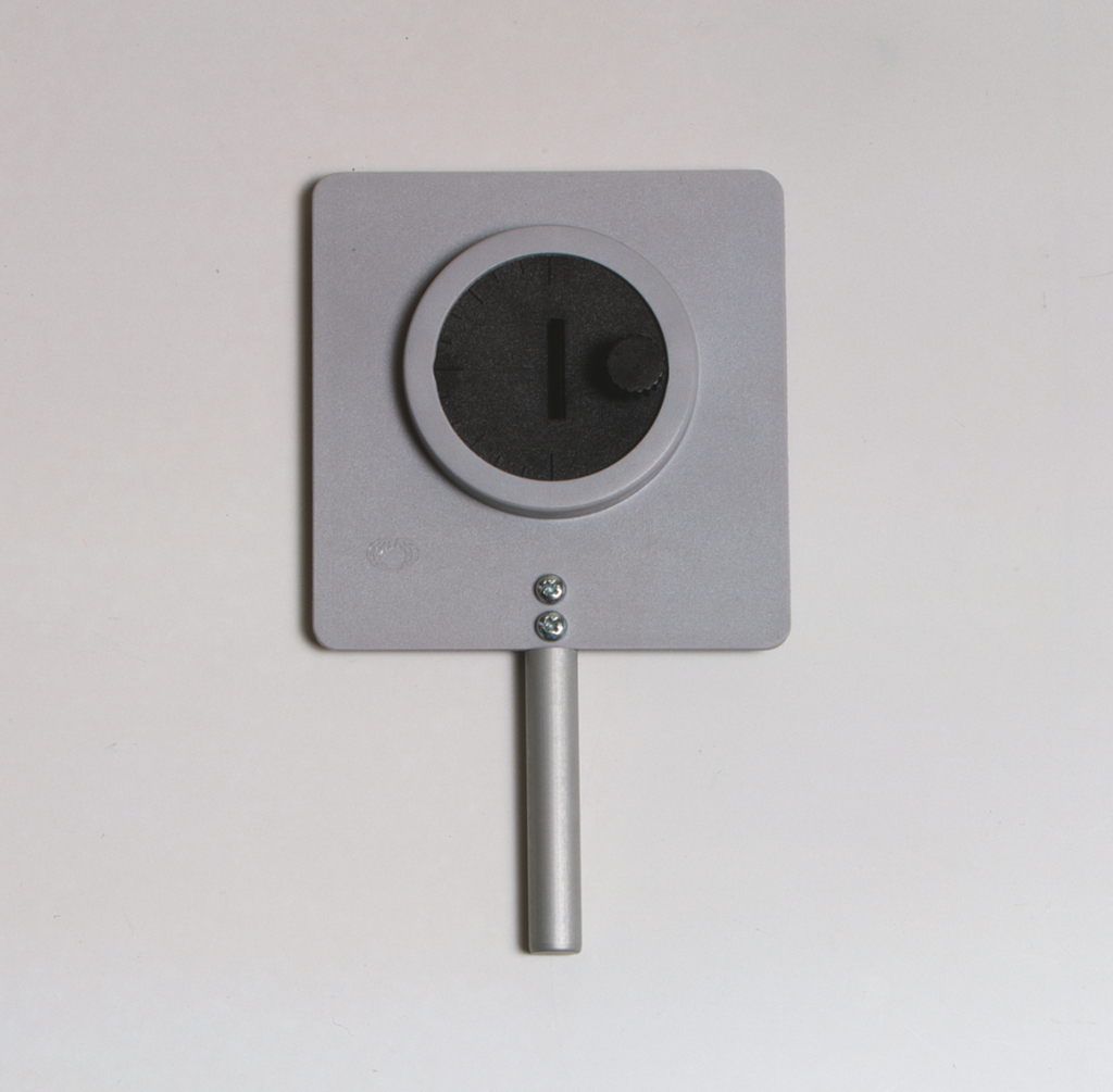 Das Bild zeigt ein Experimentiergerät, bestehend aus einer rechteckigen Platte mit einer runden Öffnung in der Mitte und einem drehbaren Regler. Es ist mit einem Metallstab ausgestattet, der als Halterung dient, und wird vermutlich zur Durchführung von physikalischen Experimenten verwendet.