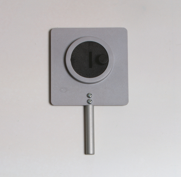 Das Bild zeigt ein Experimentiergerät, bestehend aus einer rechteckigen Platte mit einer runden Öffnung in der Mitte und einem drehbaren Regler. Es ist mit einem Metallstab ausgestattet, der als Halterung dient, und wird vermutlich zur Durchführung von physikalischen Experimenten verwendet.