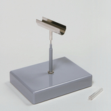 Das Bild zeigt ein Experimentiergerät, bestehend aus einer stabilen grauen Basis mit einem aufrechten Metallarm und einer schmalen Metallplatte oben. Daneben liegt ein durchsichtiges Röhrchen, das wahrscheinlich für Versuche verwendet wird.