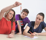 Das Bild zeigt drei Schülerinnen und Schüler, die an einem Experiment arbeiten. Eine Schülerin hält eine Spritze über einem Gerät mit Rollen, während die anderen beiden aufmerksam beobachten und Notizen machen.