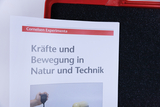 Das Bild zeigt ein Heft mit dem Titel „Kräfte und Bewegung in Natur und Technik“ von Cornelsen Experimenta. Es liegt auf einer schwarzen, schaumstoffähnlichen Unterlage und zeigt eine Hand mit einem Stoppuhrgerät.