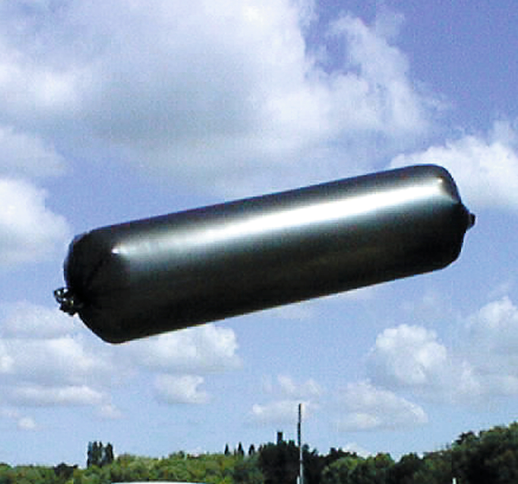 Das Bild zeigt einen großen, schwarzen, zylindrischen Ballon, der am Himmel schwebt. Die Umgebung ist eine grüne Landschaft unter einem blauen Himmel mit einigen Wolken.
