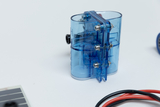 Das Bild zeigt ein transparentes, blaues Experimentiergerät mit mehreren Anschlüssen und einer Skala. Neben dem Gerät sind Kabel in Schwarz und Rot sowie ein Solarpanel sichtbar, was auf einen Zusammenhang mit technischen oder naturwissenschaftlichen Experimenten hinweist.