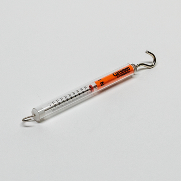 Das Bild zeigt ein Experimentiergerät, das vermutlich zur Messung von Kräften oder Gewichten verwendet wird. Es besteht aus einem transparenten Zylinder mit einer Skala und einem Haken an einem Ende, um Objekte zu wiegen oder zu testen.