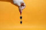 Auf dem Bild ist eine Hand zu sehen, die einen Magneten hält, unter dem zwei metallische Gewichte hängen. Der Hintergrund ist leuchtend orange, was den Fokus auf das Experimentiermaterial lenkt.
