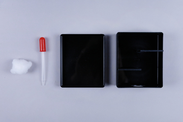 Das Bild zeigt eine Anordnung von Experimentiermaterialien, bestehend aus einem Baumwollball, einer Pipette mit rotem Kopf und zwei schwarzen Behältern. Diese Utensilien sind typisch für Schulversuche und naturwissenschaftliche Experimente.