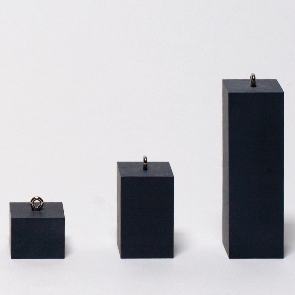 Das Bild zeigt drei schwarze, rechteckige Blöcke in unterschiedlichen Höhen, jeweils mit einer Metallöse an der Oberseite. Diese Objekte dienen vermutlich als Gewichtselemente für experimentelle Zwecke im Bildungsbereich.