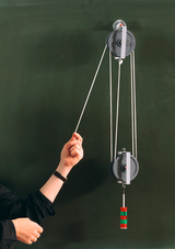 Das Bild zeigt ein Experimentiergerät zur Demonstration von Pulleys, das an einer Wand hängt. Eine Hand betätigt eine Schnur, die mit einem Gewicht verbunden ist, während ein weiteres Element sichtbar ist, das das physikalische Konzept verdeutlicht.