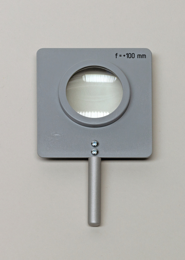 Das Bild zeigt eine graue Lupe mit einem quadratischen Rahmen und einem Griff. Die Linse hat eine Brennweite von +100 mm und ist für den Einsatz im Bildungsbereich geeignet.