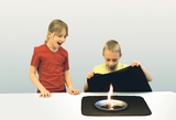 Das Bild zeigt zwei Kinder am Tisch, eines von ihnen schaut überrascht, während das andere vorsichtig einen schwarzen Stoff hebt, unter dem eine kleine Flamme auf einem Teller brennt. Die Szene vermittelt staunende Neugier und Experimentierfreude.