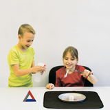 Das Bild zeigt zwei Kinder, die an einem Experiment arbeiten. Ein Junge sprüht offenbar eine Flüssigkeit auf einen Tisch, während ein Mädchen mit einer Zange ein Objekt über einer flammenden Kerze hält und lächelt.