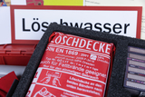 Das Bild zeigt eine Löschdecke, die in einer schützenden Schaumstoffverpackung untergebracht ist. Im Hintergrund sind weitere Materialien und ein Schild mit der Aufschrift "Löschwasser" zu sehen, die auf sicherheitsrelevante Themen im Bildungswesen hinweisen.