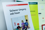 Das Bild zeigt ein Lehrmaterial mit dem Titel „Sicherer Umgang mit Feuer“ von Cornelsen Experimenta. Darunter ist eine Abbildung von Kindern, die in einem Klassenzimmer experimentieren, während auf der Tafel der Hinweis auf Sicherheit zu sehen ist.
