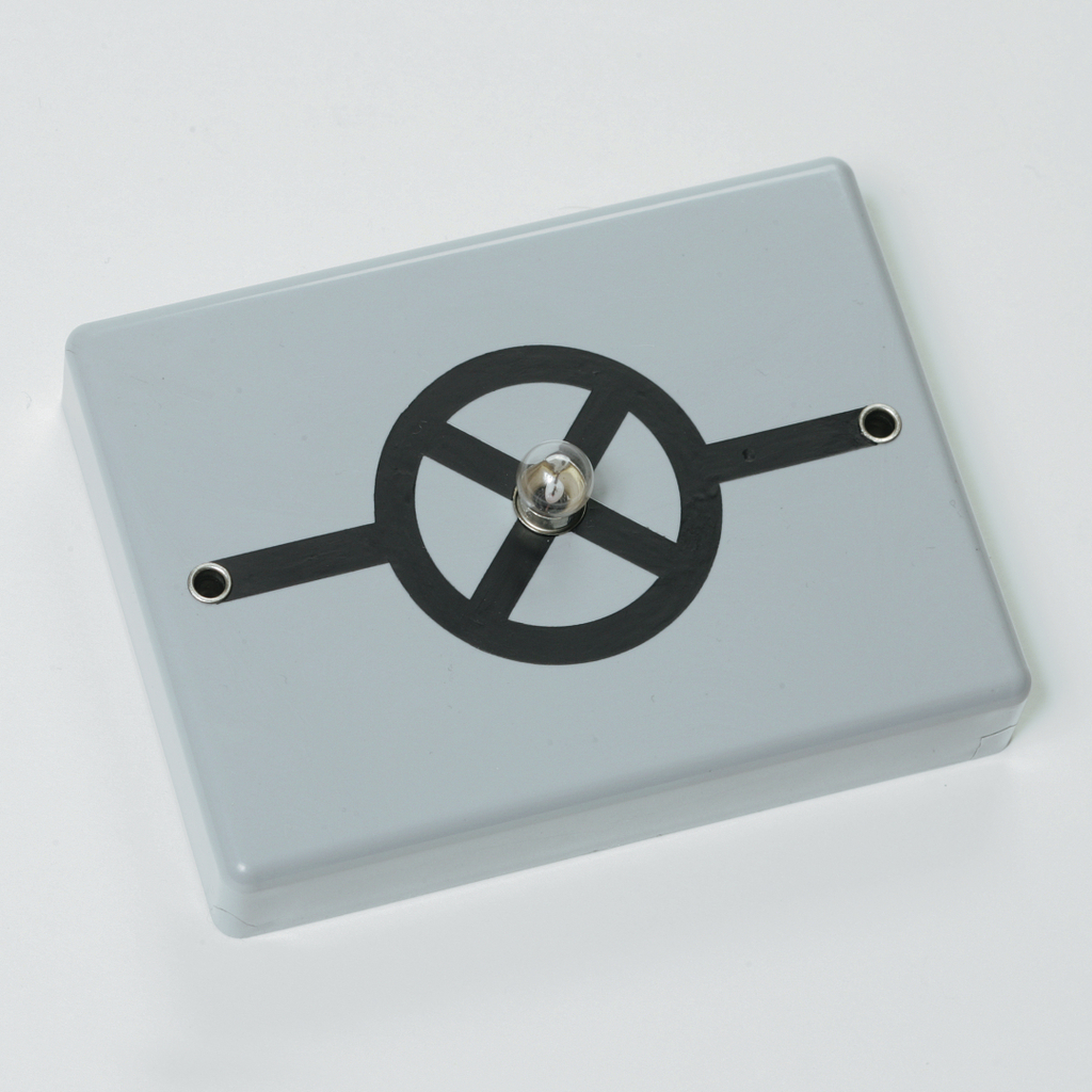 Das Bild zeigt ein elektrisches Experimentiergerät in grauer Farbe mit einer zentralen Glühbirne und einem stilisierten Schaltkreis-Design auf der Oberseite. Es ist für Bildungszwecke konzipiert und dient vermutlich zur Demonstration von elektrischen Schaltungen.