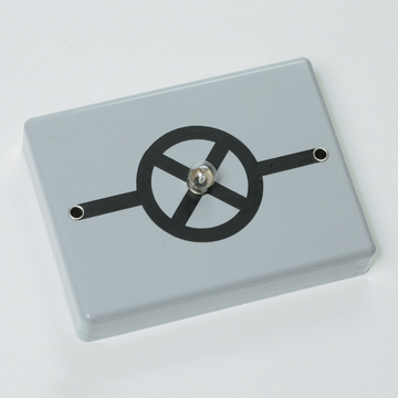 Das Bild zeigt ein elektrisches Experimentiergerät in grauer Farbe mit einer zentralen Glühbirne und einem stilisierten Schaltkreis-Design auf der Oberseite. Es ist für Bildungszwecke konzipiert und dient vermutlich zur Demonstration von elektrischen Schaltungen.