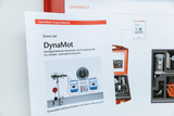 Das Bild zeigt ein Informationsblatt über das DynaMot Demo-Set, einen handbetriebenen Generator, ideal für Experimente im Schulunterricht. Im Hintergrund ist die dazugehörige Ausrüstung in einem roten Koffer abgebildet, die für Schüler- und Lehrerexperimente verwendet wird.