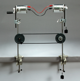 Das Bild zeigt ein Experimentiergerät mit zwei Elektromotoren, die über Riemen und Rollen miteinander verbunden sind. Es dient zur Veranschaulichung physikalischer Prinzipien, wie beispielsweise der Energieübertragung und der Bewegungsmechanik.