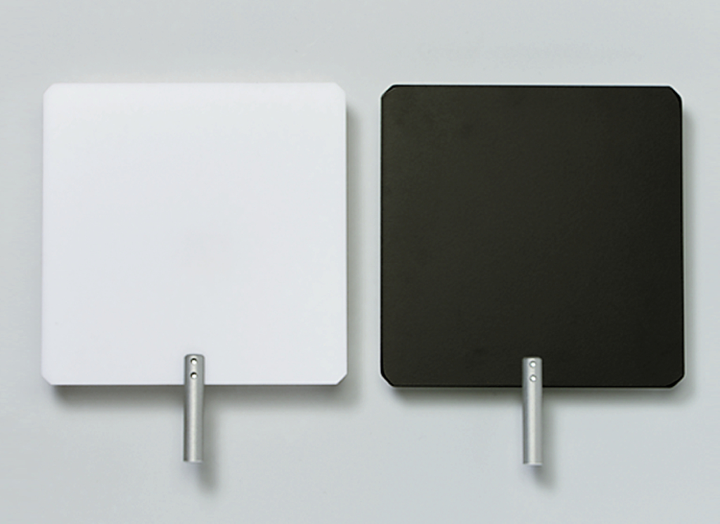 Das Bild zeigt zwei quadratische Tafeln, eine in Weiß und die andere in Schwarz, die beide einen Metallgriff an der Unterseite haben. Diese Tafeln könnten für Experimente oder Lehrzwecke im Bildungswesen verwendet werden.