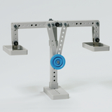 Das Bild zeigt eine einfache Wippe mit zwei Schalen an beiden Enden, die an einem stabilen Rahmen befestigt ist. In der Mitte befindet sich eine blaue Rolle, die wahrscheinlich für Experimente zur Mechanik verwendet wird.