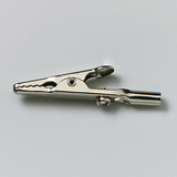 Das Bild zeigt eine metallische Krokodilklemme, die typischerweise in Experimenten verwendet wird. Sie dient zur elektrischen Verbindung von Bauteilen oder zur Befestigung von Drähten in der Wissenschaftsausbildung.