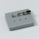 Das Bild zeigt ein graues Experimentiergerät mit zwei Metallkontakten und einem Schalter. Es dient wahrscheinlich zur Durchführung elektrischer Experimente im Bildungsbereich.