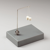 Das Bild zeigt eine einfache Hebelvorrichtung mit einem Metallbügel, der zwei kleine, weiße Gewichte trägt. Die Konstruktion steht auf einer grauen Basis und wird häufig für experimentspezifische Anwendungen im Bildungsbereich verwendet.