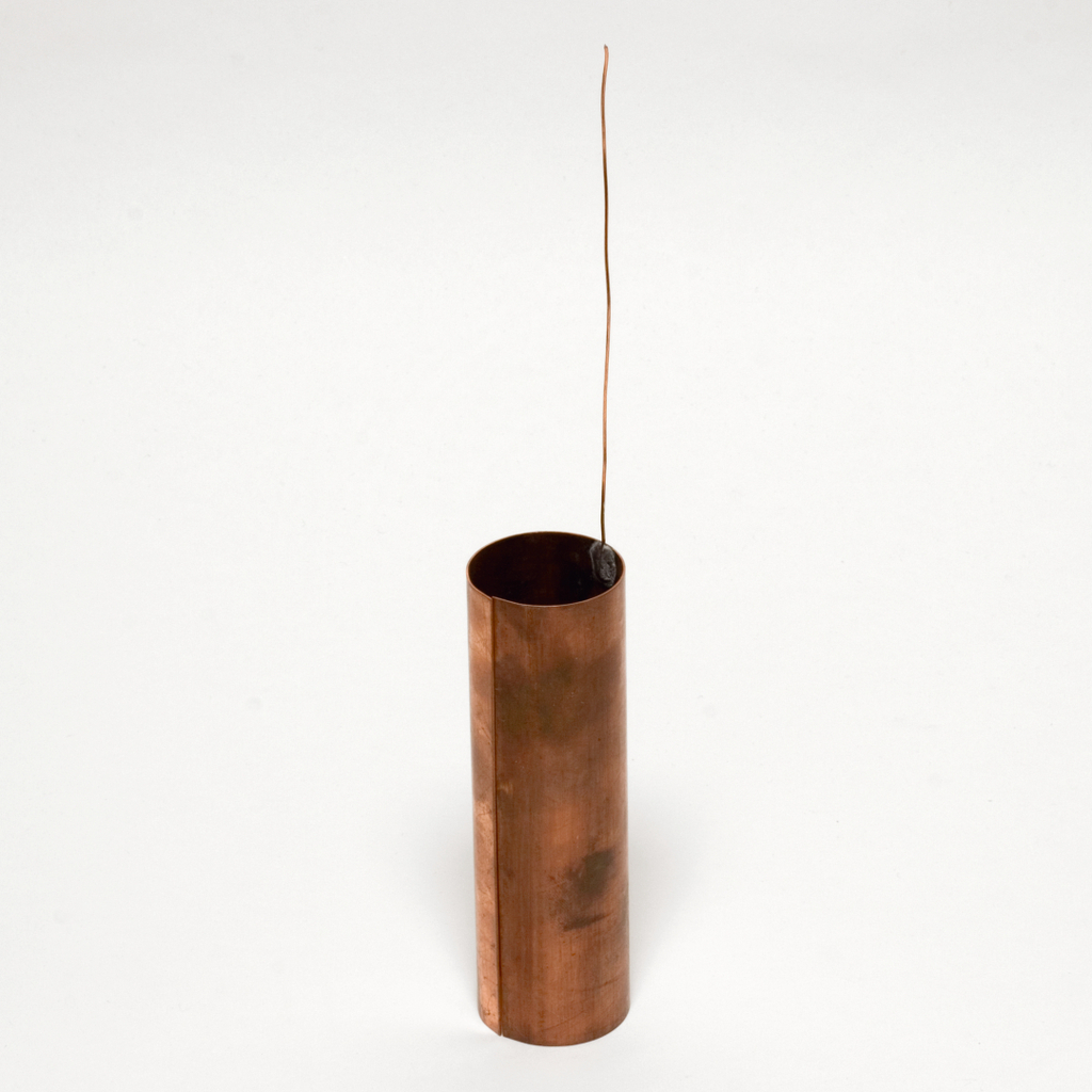 Das Bild zeigt einen zylindrischen Kupferbehälter mit einem dünnen Draht und einem kleinen schwarzen Objekt, das in der Öffnung hängt. Dieser Artikel könnte für Experimente in der Physik oder Chemie genutzt werden.