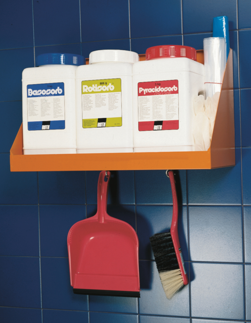 Das Bild zeigt eine orangefarbene Regalhalterung an einer blauen Wand, auf der drei Behälter mit Experimentiermaterialien stehen: Basosorb, Rotisorb und Pyracidosorb. Darunter hängen ein pinker Kehrschaufel und ein Besen, die zur Reinigung dienen.
