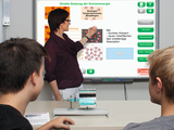 Das Bild zeigt eine Lehrerin, die an einem interaktiven Bildschirm das Thema der direkten Nutzung von Sonnenenergie erklärt. Zwei Schüler beobachten den Vortrag und arbeiten an einem Experiment, das sich mit Temperatur und Absorption beschäftigt.