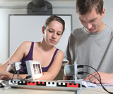 Das Bild zeigt zwei Schüler, die an einem Experimentieraufbau arbeiten. Sie verwenden verschiedene Geräte zur Durchführung eines Experiments, wobei die Schülerin den Aufbau fokussiert und der Schüler eine Anleitung studiert.