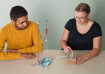 Das Bild zeigt zwei Personen, die an einem Experimentieraufbau arbeiten. Eine Person beobachtet eine von einem Stativ hängende Masse, während die andere einen Behälter mit Wasser berührt, verbunden durch einen Schlauch, der Teil des Versuchs ist.