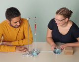Auf dem Bild sind zwei Personen zu sehen, die an einem Experiment mit einer Apparatur arbeiten, die Wasser und eine Messskala umfasst. Sie lachen und scheinen konzentriert auf die Durchführung des Experiments zu sein, das vermutlich physikalische Eigenschaften untersucht.