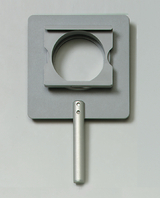 Das Bild zeigt ein Experimentiergerät mit einem grauen, rechteckigen Basisstück und einem runden Ausschnitt in der Mitte. An der Seite befindet sich ein silberner Griff, der das Halten und Hantieren des Geräts erleichtert.