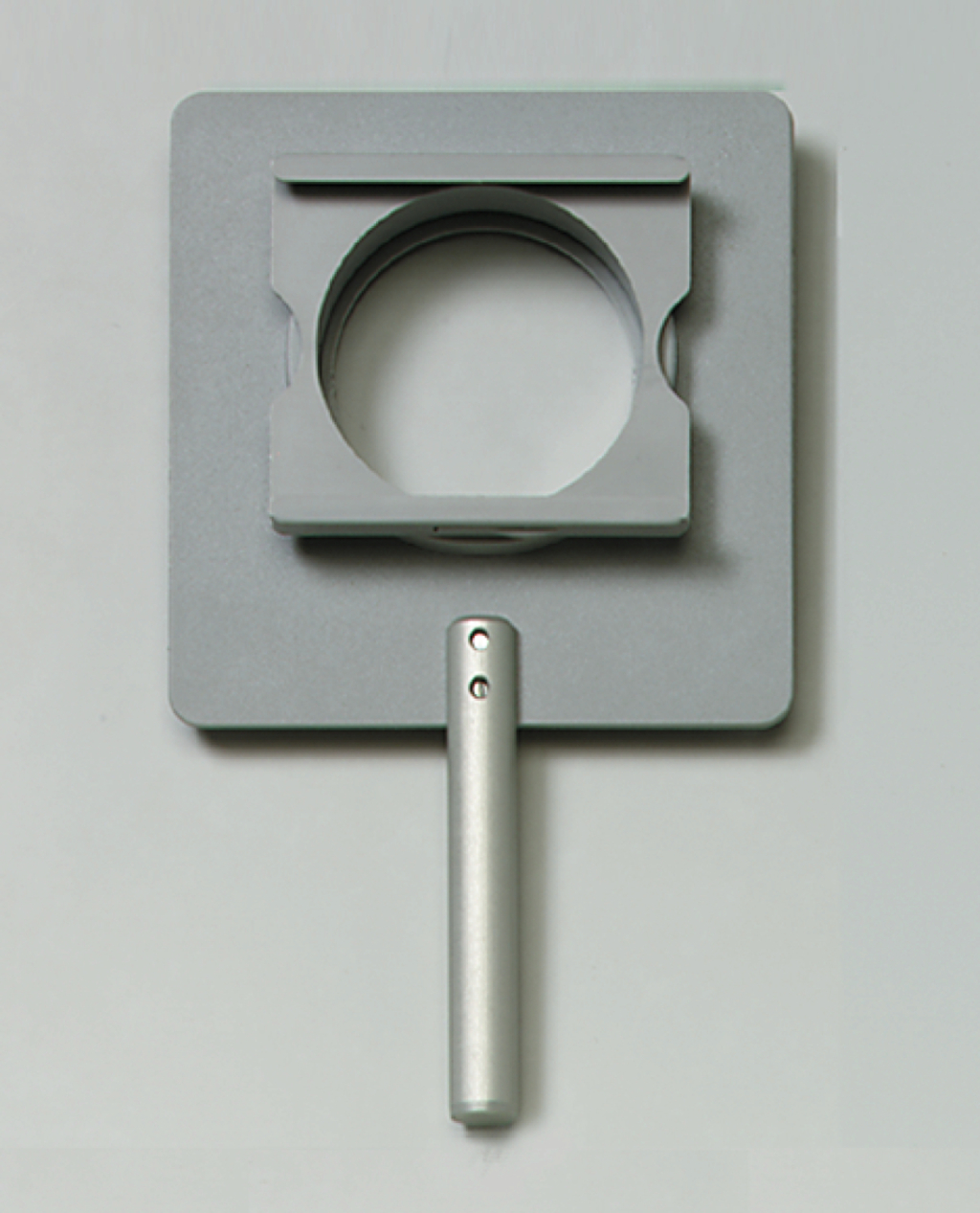 Das Bild zeigt ein Experimentiergerät mit einem grauen, rechteckigen Basisstück und einem runden Ausschnitt in der Mitte. An der Seite befindet sich ein silberner Griff, der das Halten und Hantieren des Geräts erleichtert.