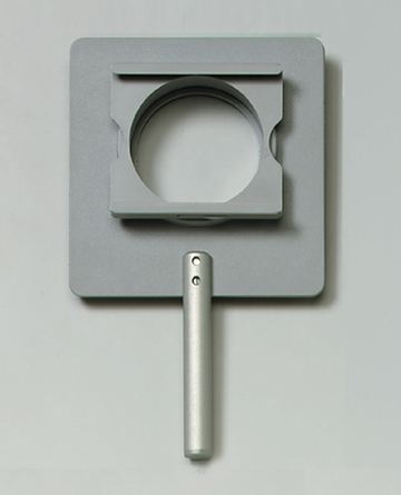 Das Bild zeigt ein Experimentiergerät mit einem grauen, rechteckigen Basisstück und einem runden Ausschnitt in der Mitte. An der Seite befindet sich ein silberner Griff, der das Halten und Hantieren des Geräts erleichtert.