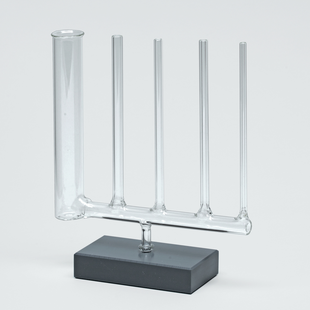 Das Bild zeigt ein experimentelles Glasgerät, bestehend aus einem Hauptrohr und vier seitlichen Röhren, das auf einem stabilen grauen Sockel montiert ist. Es wird häufig in chemischen Experimenten verwendet, um verschiedene Substanzen zu mischen oder zu beobachten.