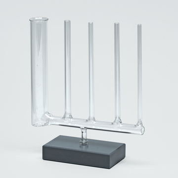 Das Bild zeigt ein experimentelles Glasgerät, bestehend aus einem Hauptrohr und vier seitlichen Röhren, das auf einem stabilen grauen Sockel montiert ist. Es wird häufig in chemischen Experimenten verwendet, um verschiedene Substanzen zu mischen oder zu beobachten.