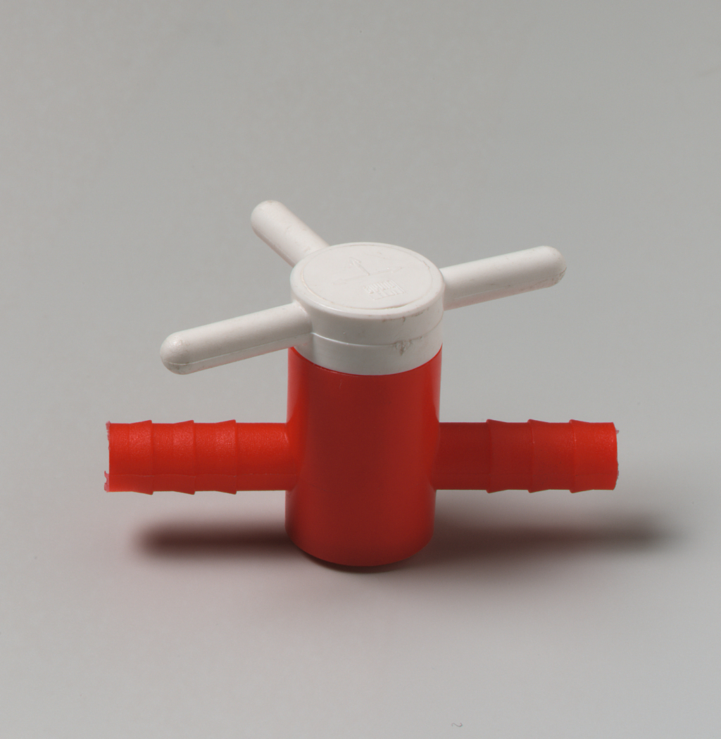 Das Bild zeigt ein rot-weißes Kunststoffteil, das anscheinend ein Ventil oder eine Verzweigung für Schläuche ist. Es hat zwei seitliche Anschlüsse und einen drehbaren Griff, um den Durchfluss zu steuern.