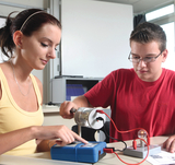 Das Bild zeigt zwei Schüler, die ein Experiment mit einer elektrischen Schaltung durchführen. Die Schülerin bedient ein Messgerät, während der Schüler eine Verbindung zu einer Glühbirne herstellt, um Funktionsweisen von Elektrizität zu erkunden.