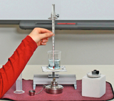 Das Bild zeigt eine experimentelle Einrichtung mit einem Becherglas, einem Thermometer und einem Heizgerät auf einem Tisch. Eine Hand hält das Thermometer in ein Becherglas mit Wasser, während eine graue Unterlage und andere Laborausrüstungen in der Nähe platziert sind.