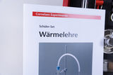 Das Bild zeigt eine Anleitung für ein Schüler-Set zur Wärmelehre aus dem Sortiment von Cornelsen. Im Hintergrund sind verschiedene Laborgeräte zu erkennen, die für Experimente in der thermo-dynamischen Forschung verwendet werden können.