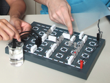 Das Bild zeigt eine Experimentierplatte mit verschiedenen elektrischen Bauteilen und einer Flüssigkeitsquelle. Zwei Personen manipulieren das Setup, um elektrische Verbindungen herzustellen und möglicherweise ein Experiment durchzuführen.